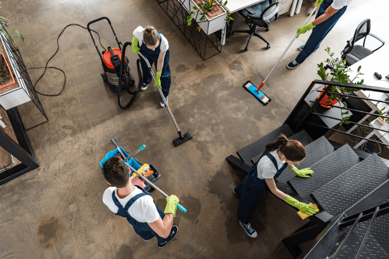 Three people cleaning a room with a concrete floor and a large window