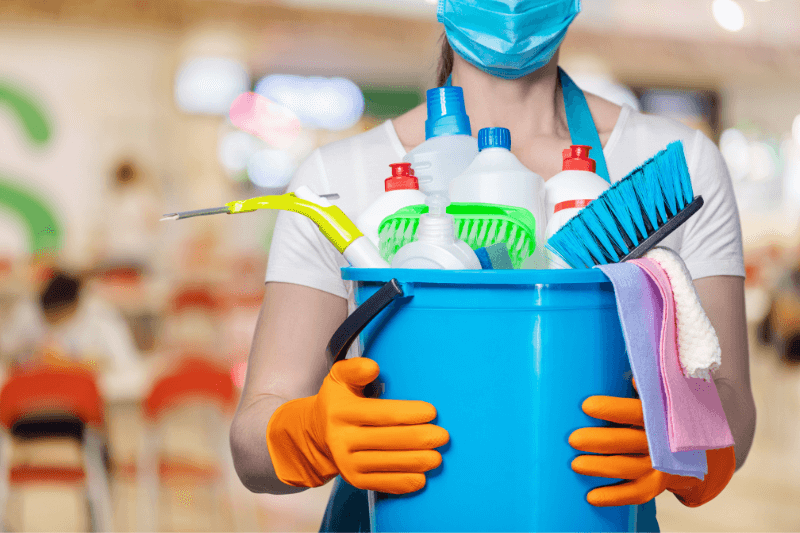 A person wearing a blue surgical mask and orange gloves holding a blue bucket filled with cleaning supplies