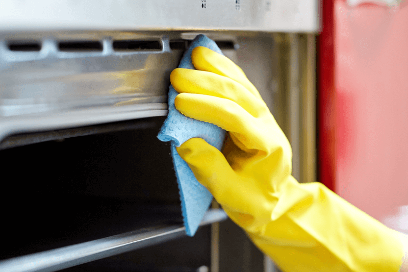 A hand wearing a yellow rubber glove cleaning the inside of an oven with a blue sponge